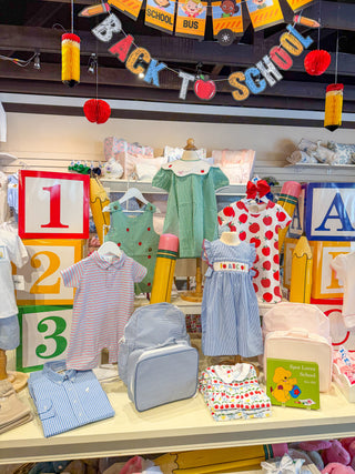 A retail display of children's clothing and accessories themed around 'Back to School', featuring colorful dresses, striped shirts, a light blue backpack, a pink lunch bag, and a children's back to school book, decorated with chalkboard-style numbers and school supplies.