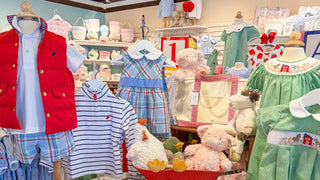 A children's clothing display in a boutique featuring multiple outfits on mannequins and tables, including striped shirts, plaid shorts, dresses, folded tops, and stuffed animals. The store interior has wooden ceiling beams, track lighting, and a colorful decorative banner overhead.