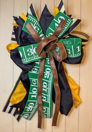 Hand-tied decorative bow featuring green football field ribbon, black and gold ribbons, black-and-white stripe ribbon, and brown faux leather ribbon.