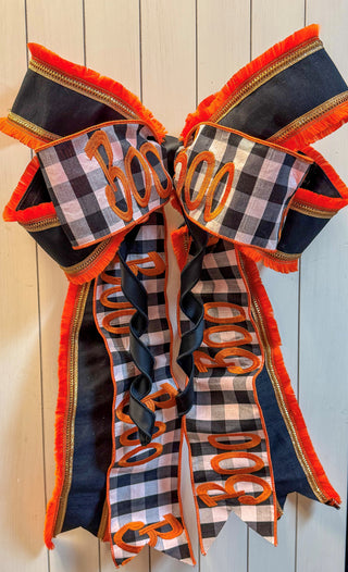 A large decorative bow with fringe and long ribbon tails, featuring a black-and-white buffalo plaid pattern with orange trim and orange script text reading '300'. The bow is photographed against a light wood plank background.