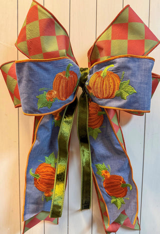 A large decorative bow with autumn-themed fabric panels, featuring orange pumpkins with green leaves on a blue background, red and green checkered sections, and a shiny olive green ribbon center.